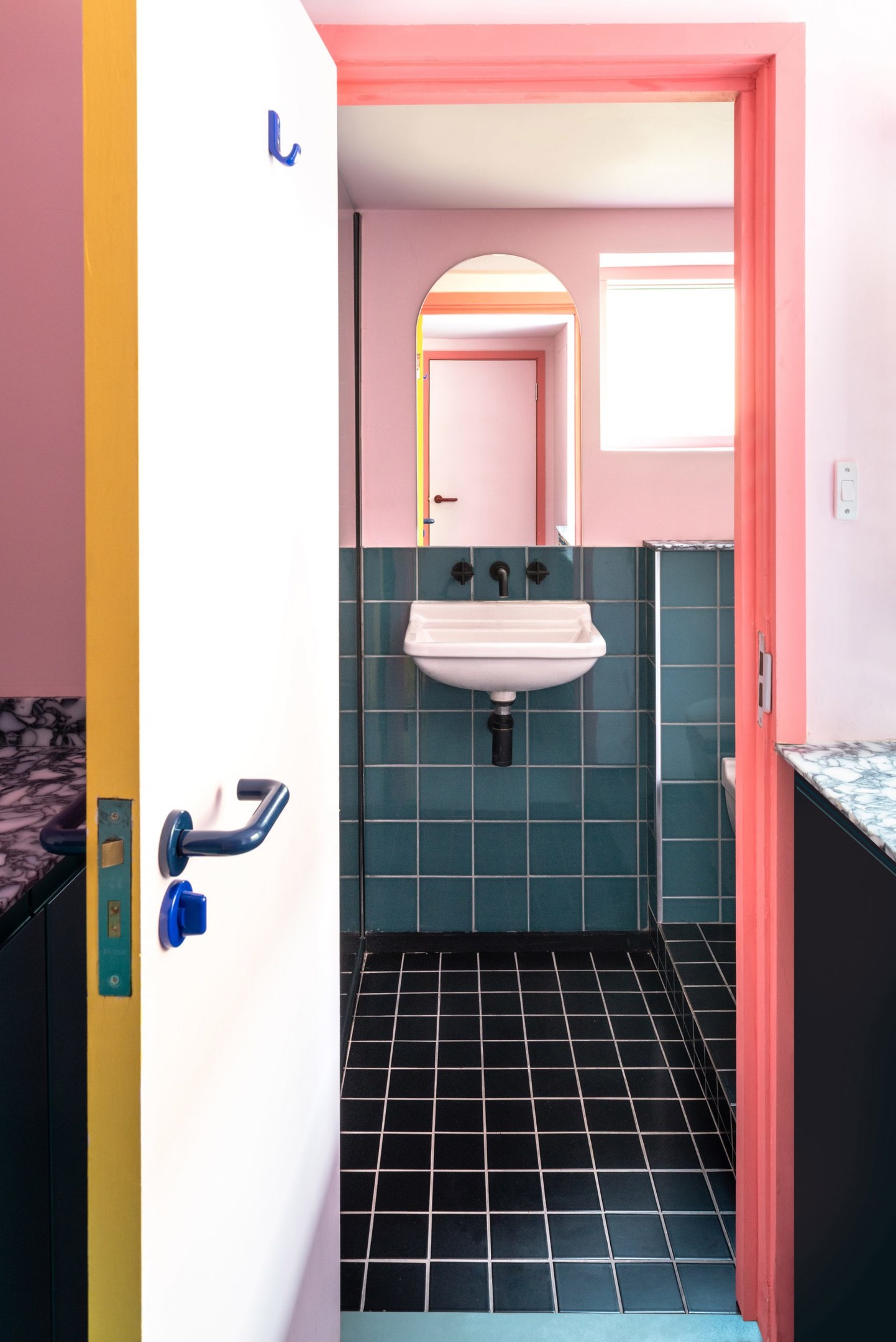 Sàn phòng tắm ốp gạch màu đen, tạo sự tương phản mạnh mẽ với nền tường hồng phối cùng gạch màu xanh. Tấm gương kiểu mái vòm phía trên bồn rửa cũng góp phần 'cơi nới' không gian và phản chiếu ánh sáng cho nhà vệ sinh nhỏ.