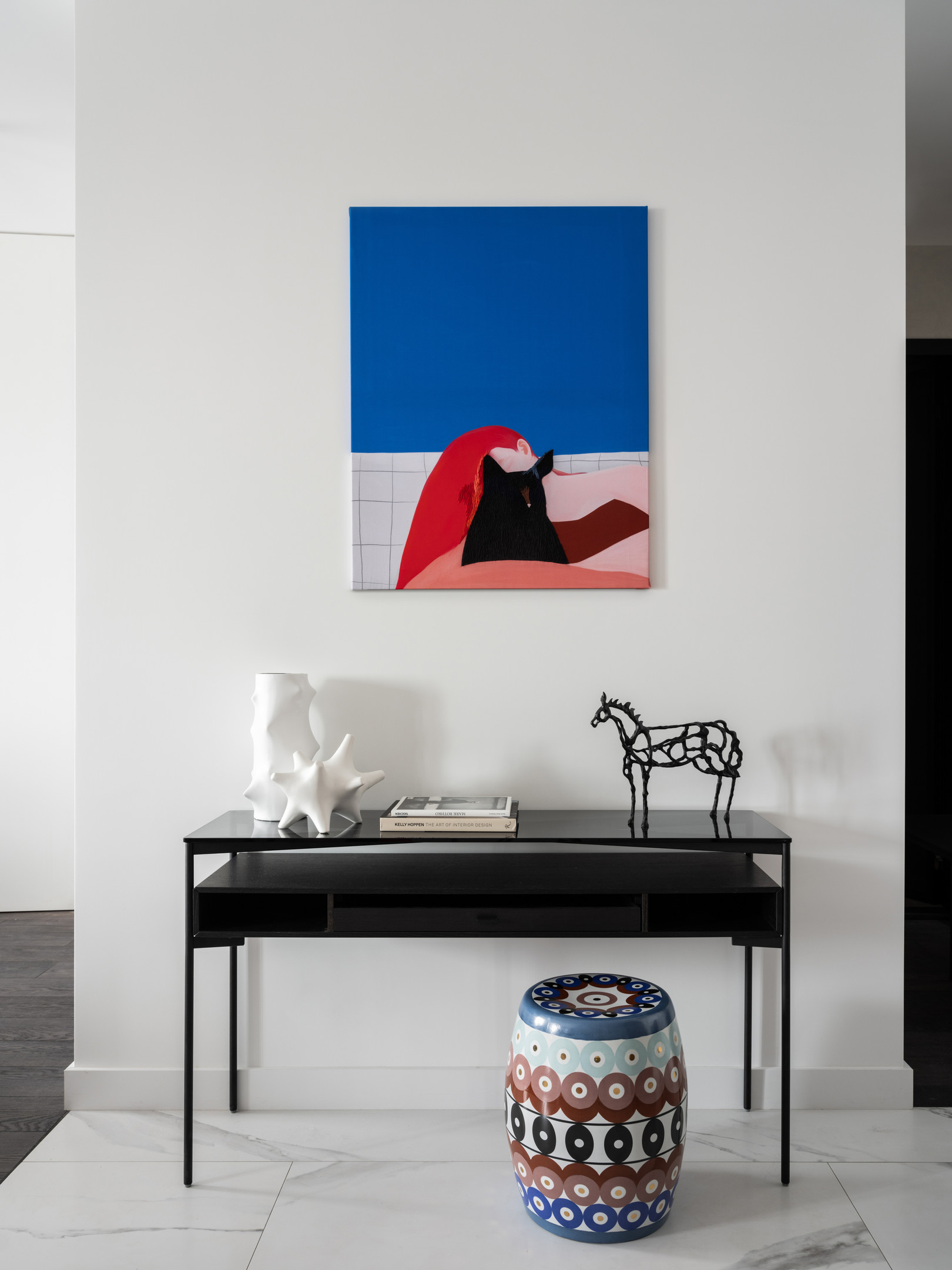 Hành lang lối vào căn hộ được bố trí một chiếc bàn màu đen để trưng bày sách và phụ kiện, nổi bật một chiếc ghế đôn như chiếc trống nhiều màu nổi bật. Trên tường là bức tranh cô gái và chú mèo đen của họa sĩ Sasha Fortova.