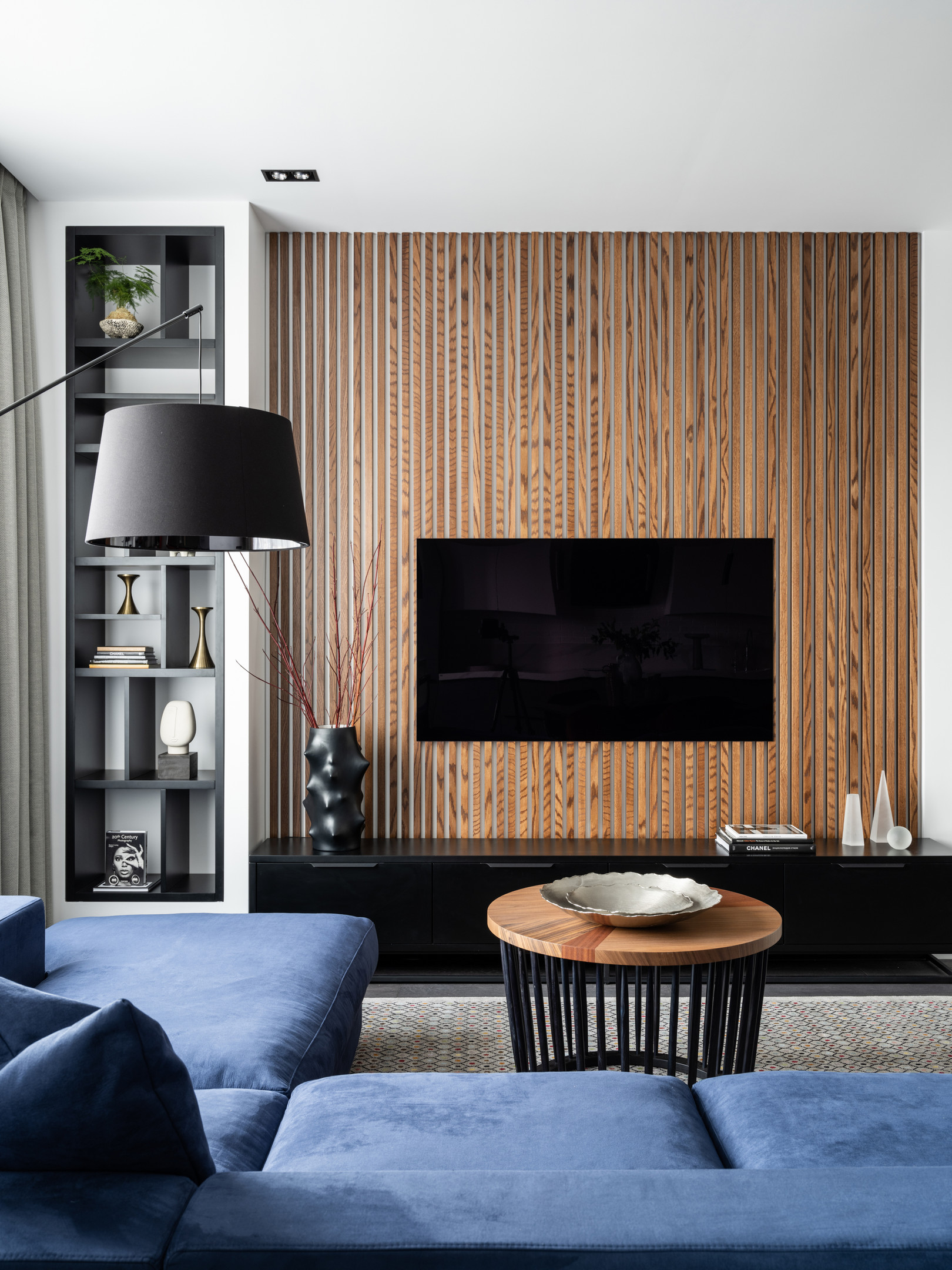 Đối diện sofa là bức tường treo tivi, được trang trí bằng những thanh lam gỗ vững chãi, cho vẻ đẹp sang trọng, đồng thời tạo sự kết nối về màu sắc với bàn nước thanh lịch. Một chiếc đèn sàn màu đen tại sofa phục vụ cho nhu cầu đọc sách.
