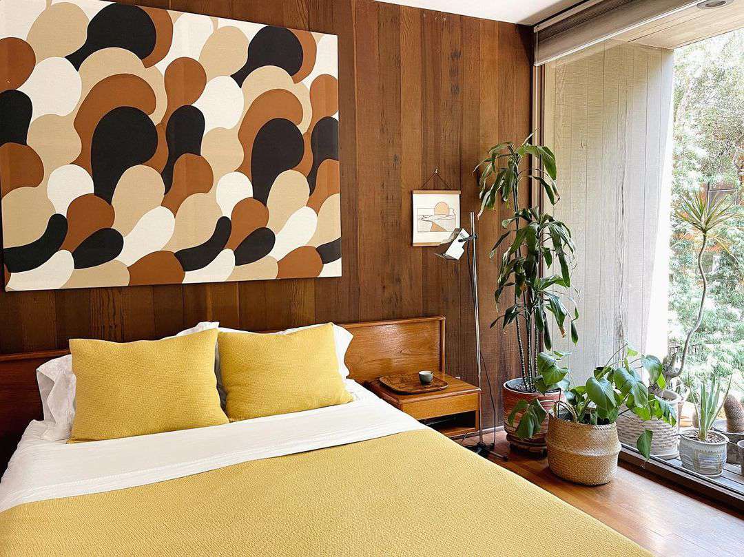 Màu nâu trầm ấm của gỗ ốp tường, vẻ độc đáo tranh trừu tượng cùng bộ chăn ga gối màu vàng khiến phòng ngủ trở nên ấm áp. Thêm vào đó là sự kết nối với khu vườn bên ngoài nhờ những chậu cảnh tươi xanh và tường kính trong suốt.