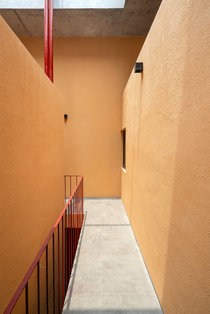 Hành lang ngôi nhà lát gạch xám thô mộc, tạo sự tương phản nhẹ nhàng với sơn tường màu cam gạch và những lan can với những chấn song kim loại màu đỏ nổi bật.