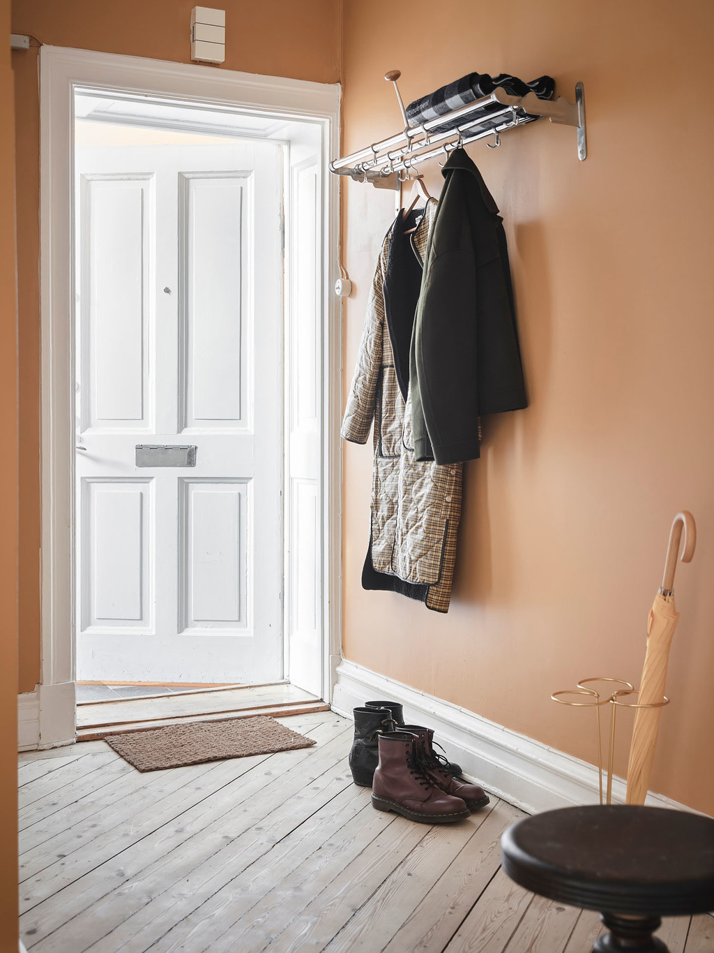 Lối vào căn hộ được sơn mới với gam màu trắng cho cánh cửa và màu cam gạch trên nền tường tươi sáng và ấm áp. Giá treo quần áo cùng với giá đỡ những chiếc ô bằng kim loại sơn mạ vàng cho lối vào gọn đẹp.