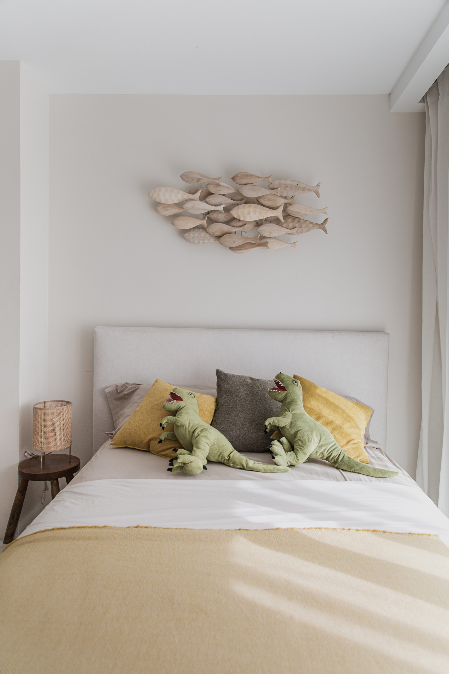 Đầu giường trang trí một đàn cá gỗ trông rất sinh động. Trên giường còn có cả 2 chú thú bông hình khủng long màu xanh đáng yêu. Chiếc đèn ngủ bằng mây tre đan cũng đi theo phong cách sử dụng vật liệu tự nhiên khi lựa chọn nội thất.