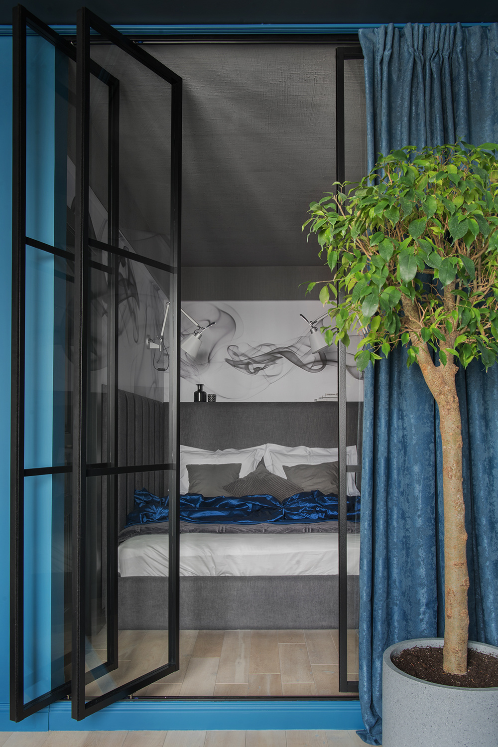 NTK nội thất Dina Vafina đã 'đóng khung' phòng ngủ nhỏ gọn này bằng cửa kính khung màu đen cổ điển. Thêm vào đó, sự kết hợp màu xám sang trọng với màu xanh lam yên tĩnh cùng chậu cây cảnh tươi xanh cho phòng ngủ thêm thư giãn.