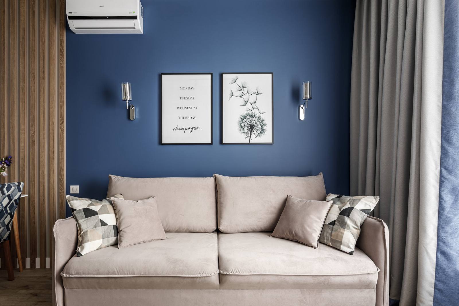 Phòng khách với duy nhất chiếc ghế sofa êm ái màu be đậm, phía sau bức tường sơn màu xanh lam đậm là 2 bức tranh nhẹ nhàng cùng đèn gắn tường cân đối, hài hòa. Việc loại bỏ hoàn toàn chiếc bàn nước tạo nên khoảng trống rộng rãi ở trung tâm.