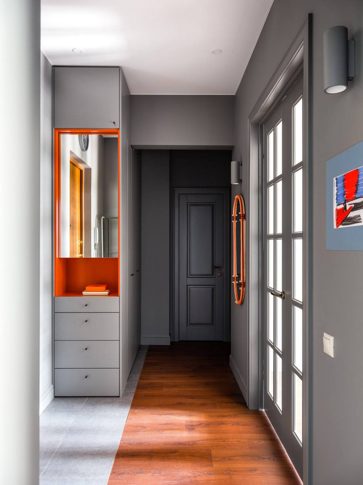 Lối vào căn hộ được thiết kế đơn giản nhưng sang trọng với sơn tường màu xám đậm, kết hợp tủ lưu trữ ốp gương với hộc tủ màu cam bắt mắt, góc phải còn có tấm gương hình oval với viền khung đồng sắc ấn tượng.