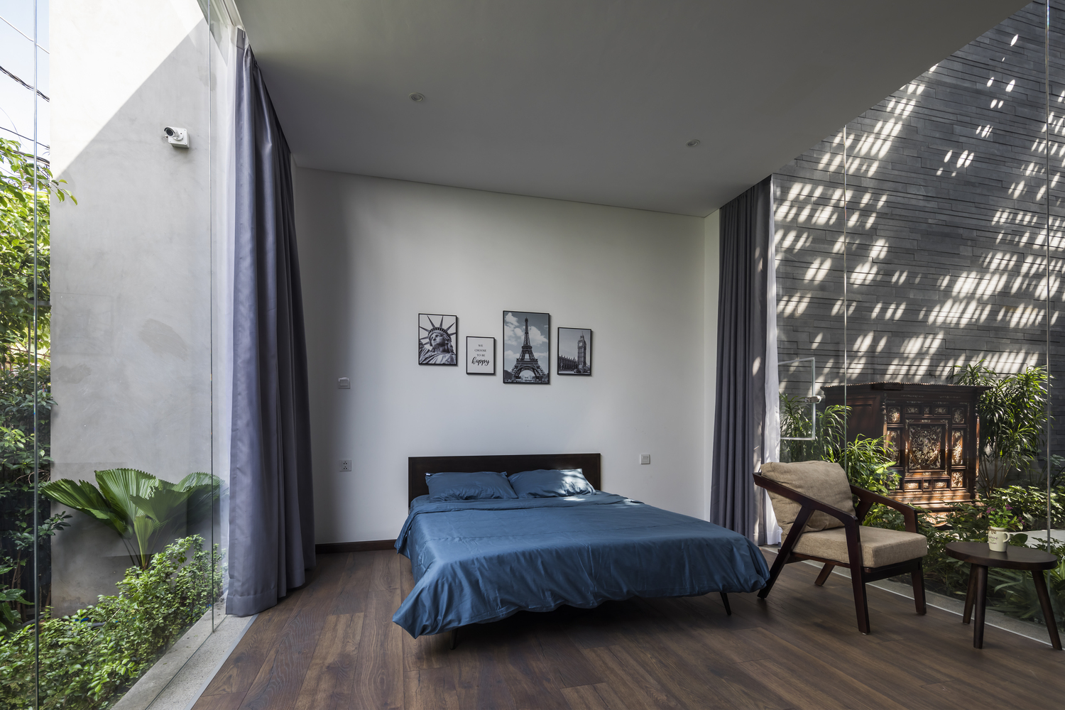 Một phòng ngủ khác được bố trí ở tầng trệt, với cửa kính trong suốt nhìn ra cây cối xanh mướt, kèm theo rèm che để tạo sự riêng tư khi cần thiết.