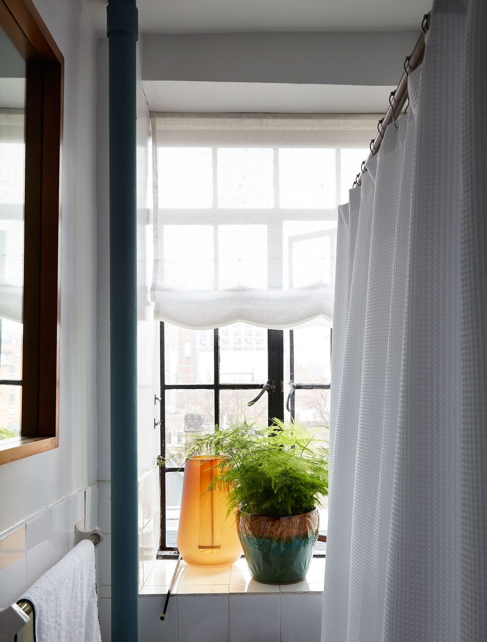 Những chiếc bình thủy tinh màu cam sáng và chậu cây bằng gốm sứ bóng mượt làm nổi bật phòng tắm nhỏ với sắc trắng chủ đạo, kết hợp với chúng là tấm rèm Roman mỏng manh cổ điển cho cửa sổ thêm phần duyên dáng.