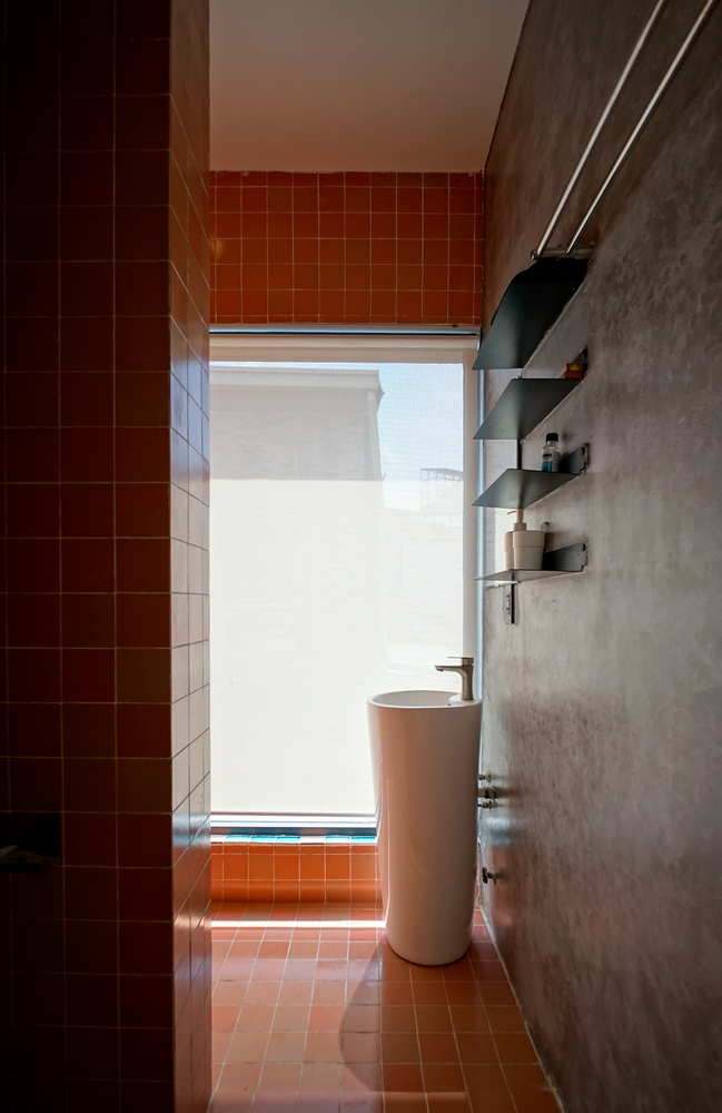 Phòng tắm và nhà vệ sinh được thiết kế với sự đối lập giữa tường xám lạnh với gạch ốp lát hình vuông màu cam để xua đi sự lạnh lẽo vốn có của căn phòng này.
