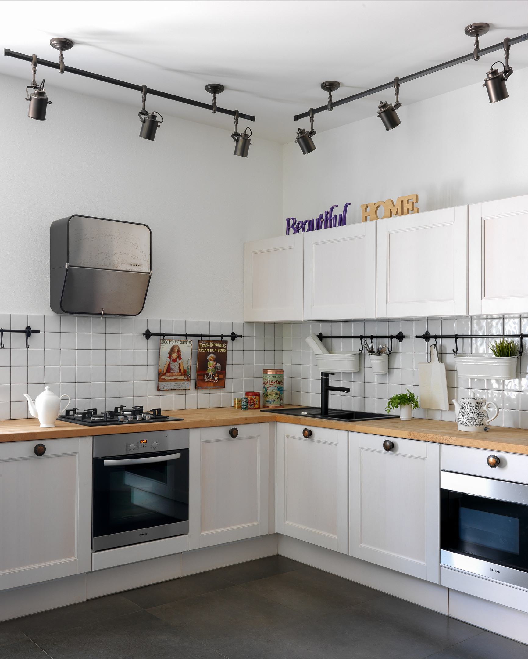 Phòng bếp thiết kế kiểu chữ L vuông góc với căn phòng tạo nên không gian nấu nướng tiện nghi. Bức tường tại khu vực backsplash được ốp gạch vuông màu trắng, kết hợp những thanh treo màu đen để trưng bày đồ dùng nhà bếp.