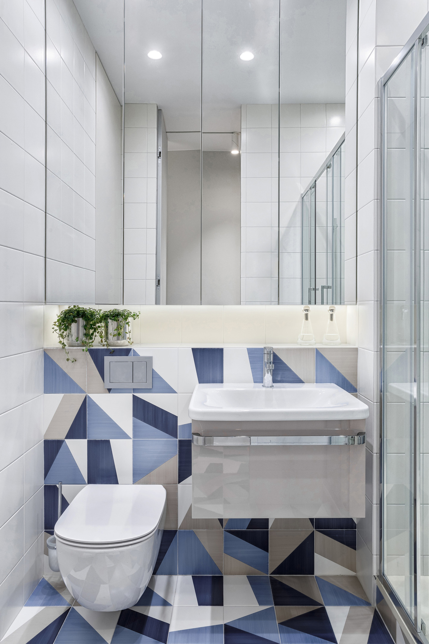 Phòng vệ sinh nhỏ gọn nhưng thoáng sáng nhờ hệ tủ lưu trữ phía trên bồn rửa tích hợp gương soi phản chiếu ánh đèn. Ngoài ra, sự kết hợp gạch mosaic trắng và gạch mosaic có màu sắc và họa tiết từ sàn đến tường cho cái nhìn sinh động hơn.
