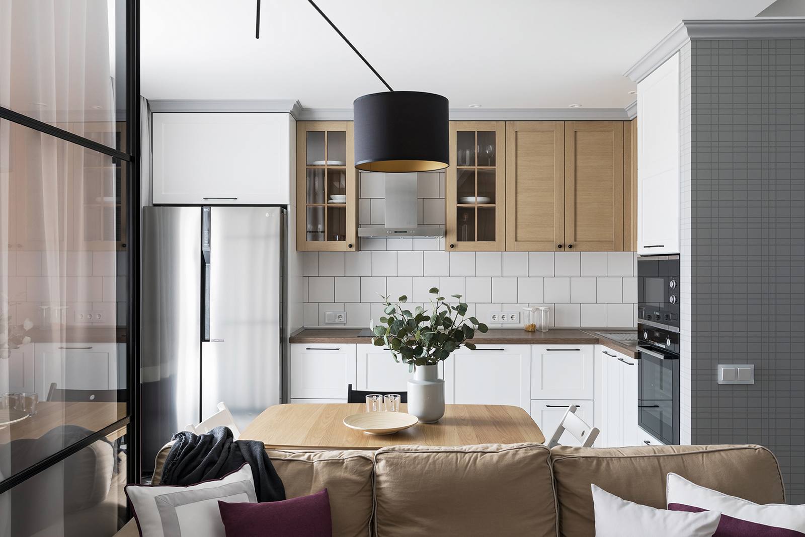 Phòng bếp thiết kế kiểu chữ L, giải pháp phù hợp cho căn hộ vừa và nhỏ. Hệ tủ bếp trên bằng gỗ tạo sự tương phản với tủ bếp dưới và backsplash ốp gạch mosaic trắng. Bề mặt tủ lạnh bằng thép không gỉ bóng bẩy, phản chiếu ánh sáng từ ô cửa đầy nắng.