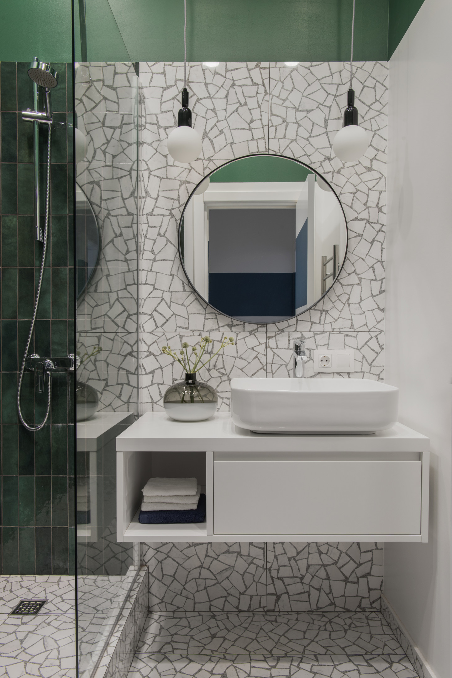 Tường và sàn phòng tắm được ốp bằng gạch họa tiết tương đồng với hành lang lối vào. Buồng tắm đứng ốp gạch thẻ dọc màu xanh ngọc lục bảo, phân vùng với tủ vanily bằng cửa kính trong suốt thoáng sáng.