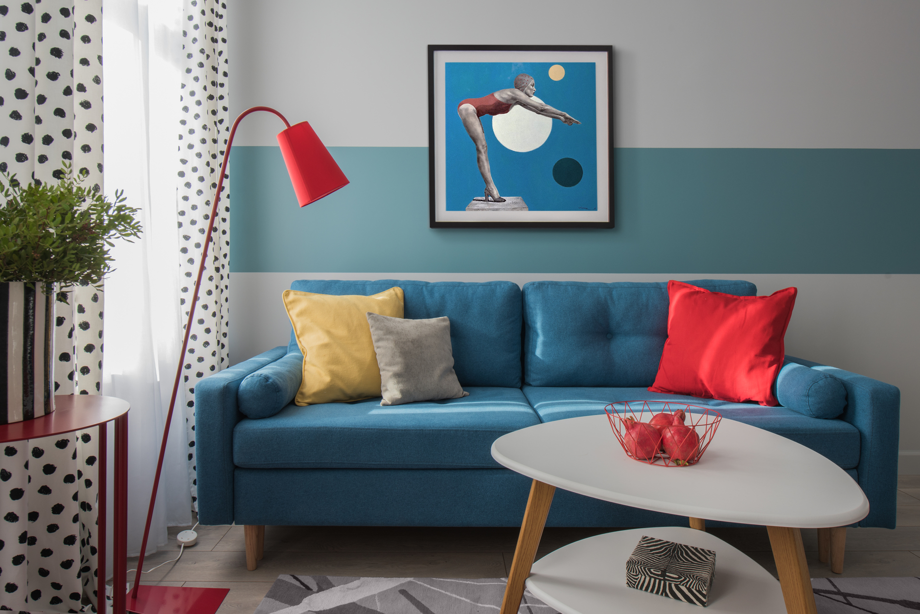 Ghế sofa xanh lam tone màu đậm hơn so với sơn sọc kẻ ngang. Những chiếc gối màu đỏ, vàng, xám cùng đèn sàn dáng cao sắc đỏ trông vừa thanh mảnh vừa linh hoạt. Bức tranh vận động viên trang trí cho tường sau sofa thêm sinh động.