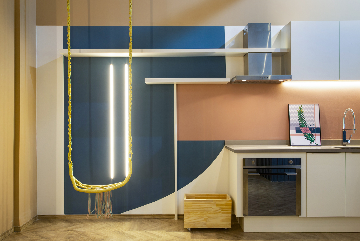 Phòng bếp thiết kế kiểu chữ I - giải pháp hoàn hảo cho căn hộ nhỏ vì tiết kiệm diện tích sàn, tối ưu hóa chiều cao trần và tạo lối đi thông thoáng. Backsplash hồng phấn cùng chiếc võng treo màu vàng tươi nổi bật trên phông nền trắng - xanh lam đậm.