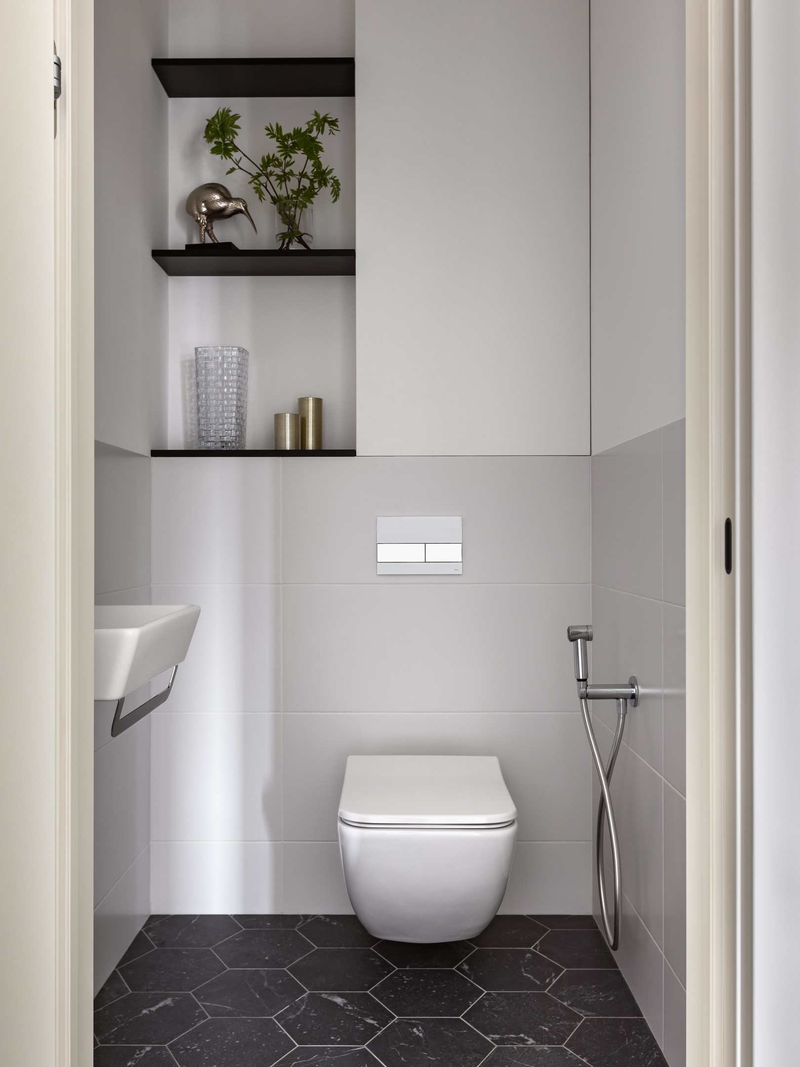Khu vực toilet được thiết kế khá tương đồng về vật liệu với lối vào, tường sơn màu trắng kết hợp gạch ốp xám nhạt, phần sàn lát gạch tổ ong. Toilet thiết kế gắn tường để giải phóng diện tích mặt sàn. Hốc tường phía trên được tận dụng để trang trí vài tác phẩm chủ nhân yêu thích.