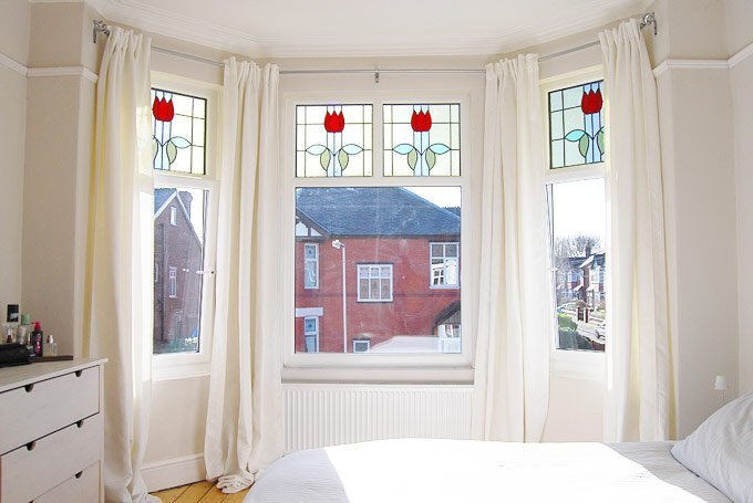 Phòng ngủ xinh xinh với tone màu trắng thanh lịch, rèm che nhẹ nhàng. Phần trên cao của các khung cửa sổ được lắp đặt cửa kính màu với hình ảnh những bông hoa hồng cách điệu duyên dáng trên phông nền trắng.