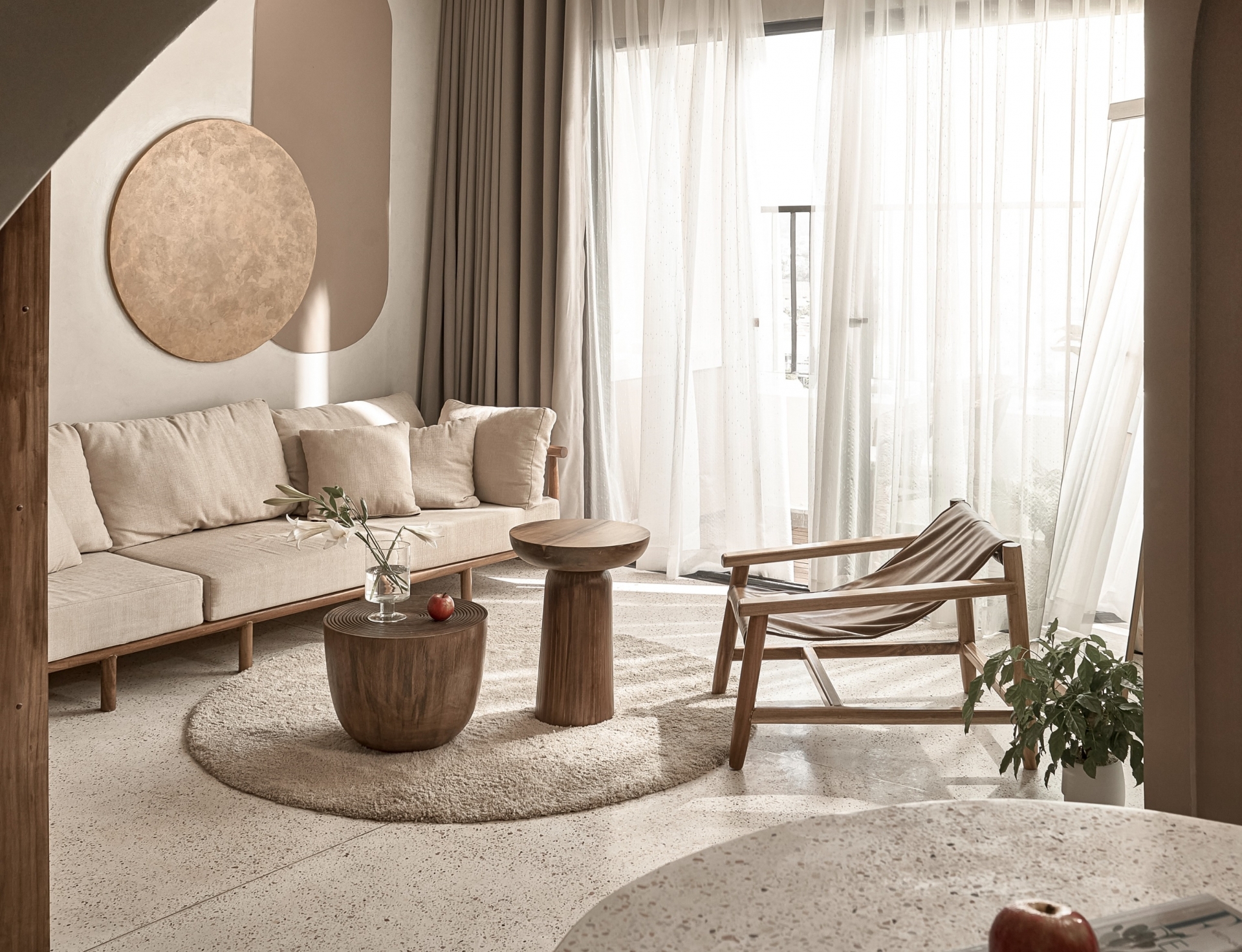 Phòng khách bố trí bên logia đầy nắng ấm với rèm che mỏng nhẹ. Ghế sofa màu be kết hợp bộ bàn nước gỗ cho vẻ đẹp mộc mạc, bố trí trên tấm thảm hình tròn êm ái.