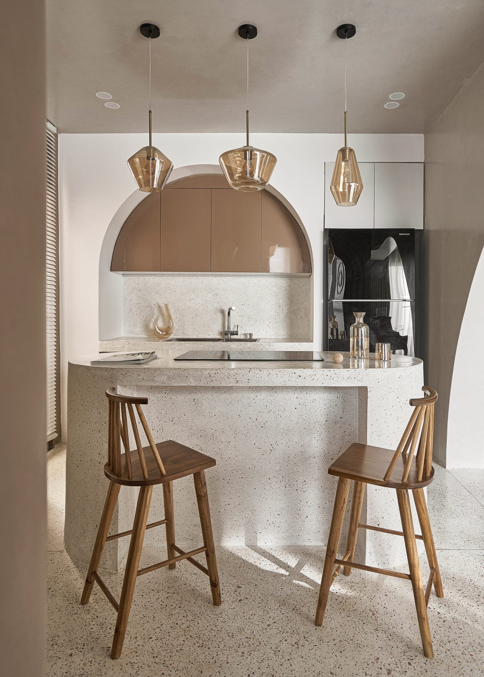 Tủ bếp màu nâu với mái vòm cong duyên dáng trên nền tường trắng. Họa tiết Terrazzo xuất hiện ở mọi nơi. Đối diện là đảo bếp, sử dụng như bàn ăn tiện lợi với bộ đèn thả trần sang chảnh.