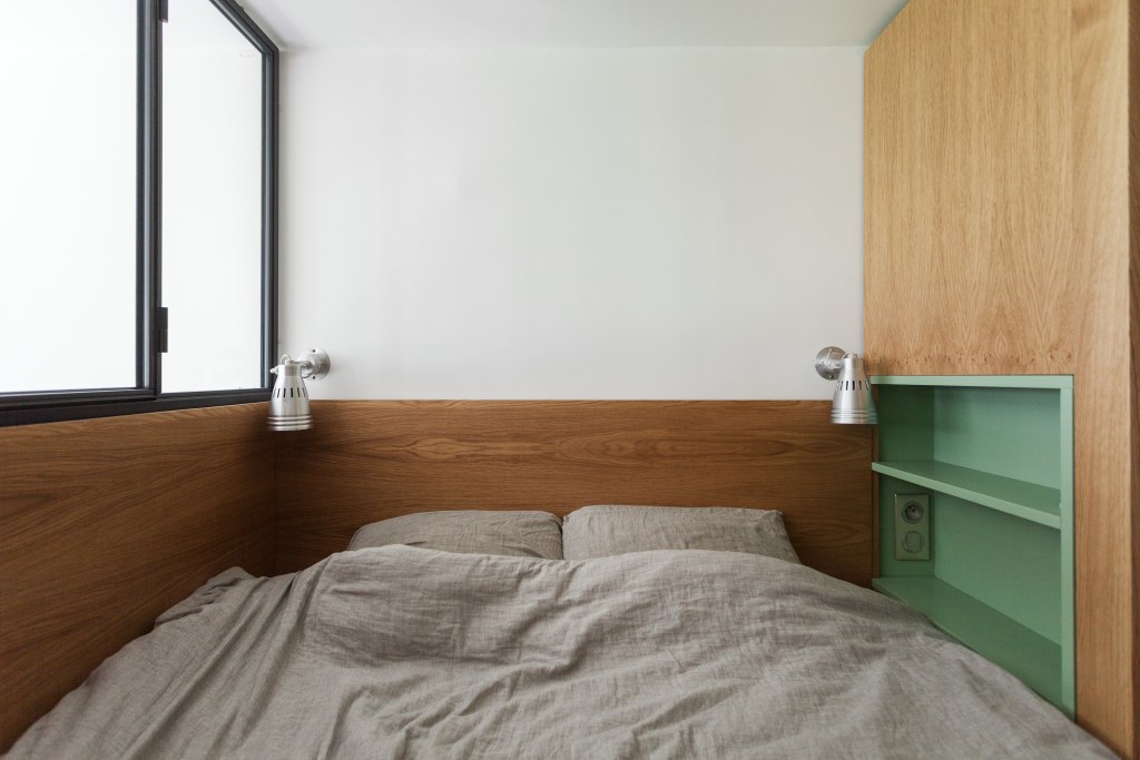 Phòng ngủ tối giản với sơn tường màu trắng kết hợp gỗ đồng màu với tổng thể căn hộ. Đèn gắn trần 'có cặp có đôi' bố trí đối xứng nhau. Thay vì sử dụng táp đầu giường, NTK đã tích hợp kệ mở sơn màu xanh lá cây ốp trong tường gỗ.
