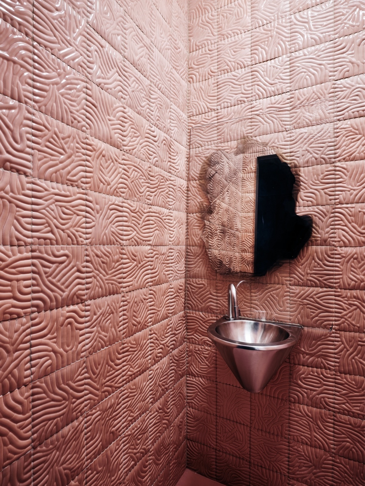 Phòng tắm được trang trí độc lạ với chiếc bồn rửa tay tựa như một chiếc phễu, bên trên là tấm gương hình thù lạ mắt. Tất cả được lắp đặt trên bức tường ốp gạch màu hồng đất họa tiết 3D sống động.