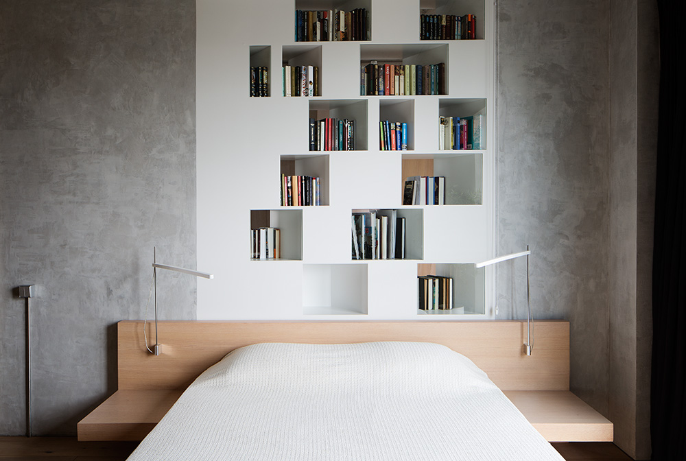 Phòng ngủ thiết kế tối giản nhưng vẫn ấn tượng nhờ bức tường bê tông xám xung quanh kệ sách âm tường màu trắng. Khu vực đầu giường bằng gỗ thiết kế 'kiêm nhiệm' chức năng của táp đầu giường tiện lợi.