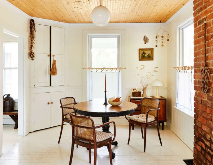 Phòng ăn với chiếc bàn tròn và ghế gỗ kết hợp mây tre đan mộc mạc. Sự tương phản giữa trần gỗ, sàn - tường trắng và một phần tường gạch thô cho không gian ấm cúng hơn.