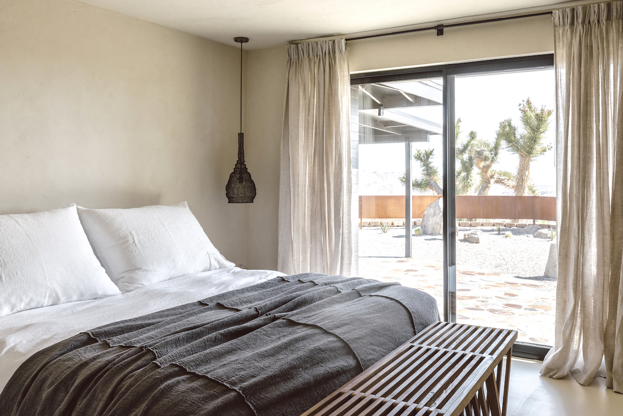 Phòng ngủ chính thiết kế theo phong cách tối giản với các tone màu trung tính như nền tường và rèm vải màu be, bộ chăn ga gối trắng - xám và băng ghế cuối giường bằng gỗ. Từ phòng ngủ, bạn có thể ngắm cảnh đẹp qua khung cửa trượt trong suốt.