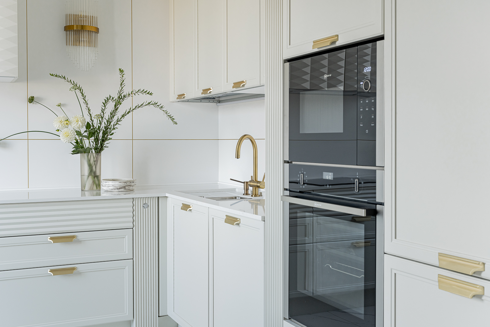 Phòng bếp tiện nghi với thiết kế kiểu chữ L, sử dụng tone màu trắng sạch sẽ, nhấn nhá các chi tiết phần cứng mạ vàng đồng và thiết bị nấu nướng màu đen sang trọng.