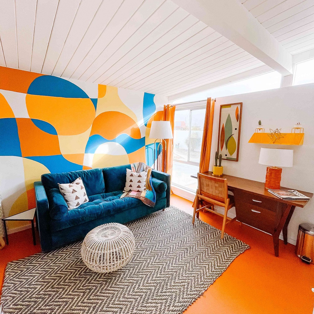 Nếu bạn thích sự rực rỡ cùng hình ảnh trừu tượng lý thú thì sẽ bị thu hút bởi phòng khách kết hợp góc làm việc tại gia này. Không chỉ là bức tường, sàn nhà cũng sử dụng tone màu cam, tương phản mạnh mẽ với trần nhà trắng sáng.