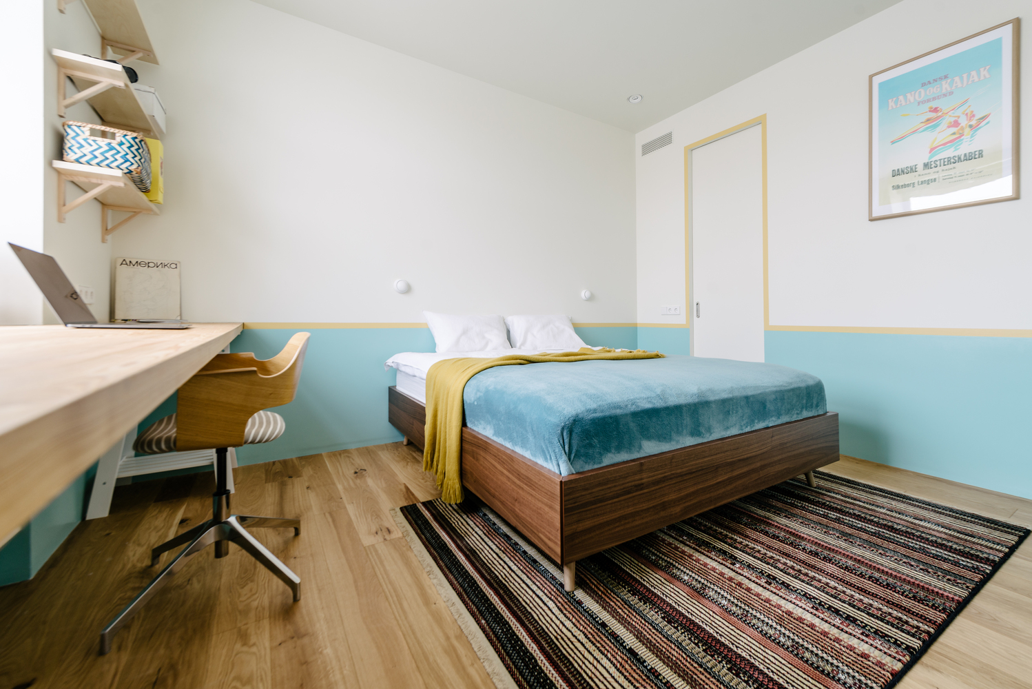Phòng ngủ sử dụng sơn tường màu trắng và xanh lam nhạt, kết hợp đường sơn viền màu vàng mù tạt như một dải phân cách nổi bật. Thảm trải sàn họa tiết kẻ sọc giúp 'nới rộng' chiều dài căn phòng bằng thị giác.