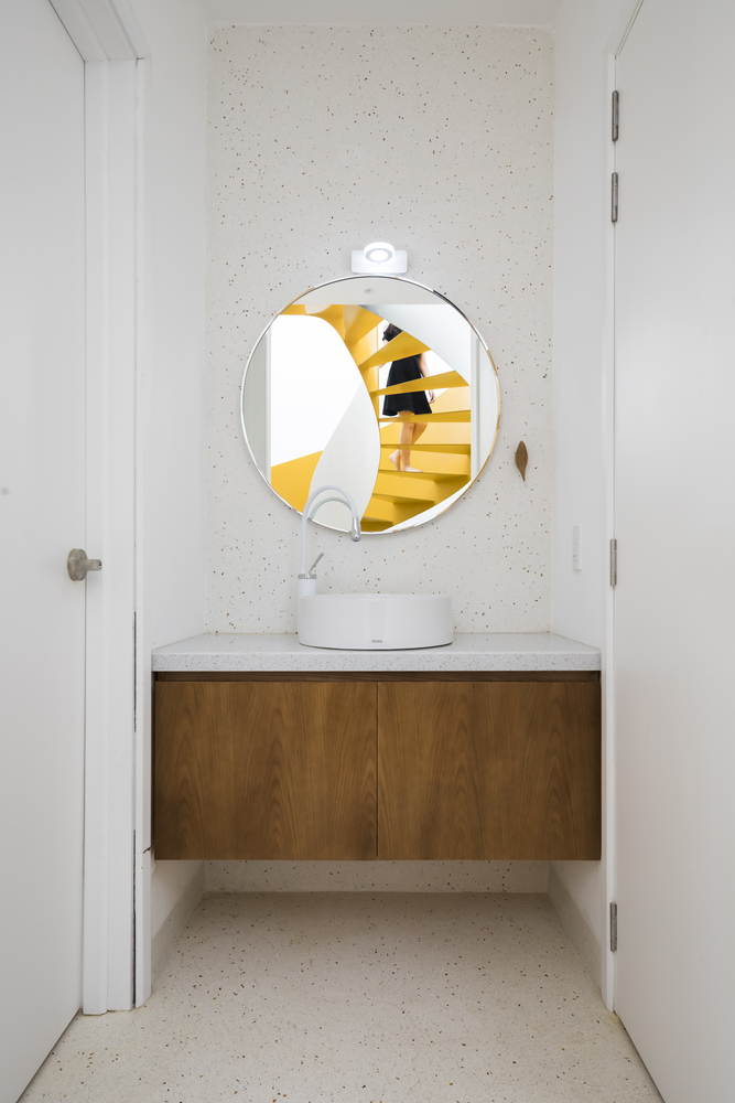 Khu vực rửa tay với chiếc tủ vanity gắn tường gọn gàng, màu gỗ trầm tạo vẻ ấm áp giữa sắc trắng. Đặc biệt, tấm gương hình tròn phản chiếu hình ảnh cầu thang xoắn màu vàng đẹp mắt.