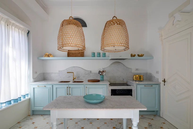 Phòng bếp mát mẻ với tone màu trắng - xanh lam, rèm che mỏng nhẹ, những chi tiết mạ vàng lấp lánh kết hợp backsplash ốp đá tự nhiên theo phong cách Địa Trung Hải.