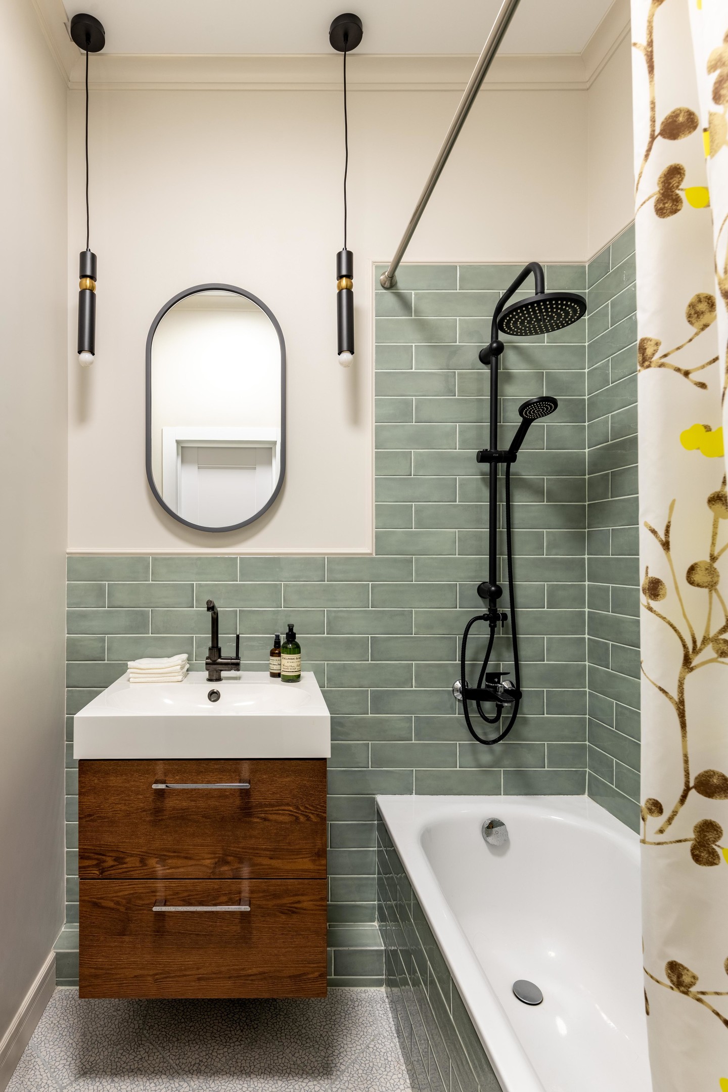 Khu vực phòng tắm phân vùng với toilet bằng rèm che vải nhẹ nhàng. Bức tường và bồn tắm ốp gạch màu xanh nhạt tươi mát. Hệ thống đèn thả trần cân xứng với gương hình oval tạo điểm nhấn cho phòng tắm nhỏ.