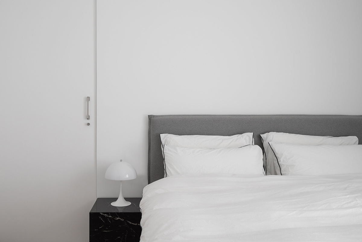 Phòng ngủ cực kỳ đơn giản với chiếc giường khung màu xám thanh lịch, chăn ga gối trắng, táp đầu giường bằng đá cẩm thạch đen tạo sự tương phản và nét sang chảnh. Hầu như không có bất cứ phụ kiện trang trí nào trong không gian thư giãn.