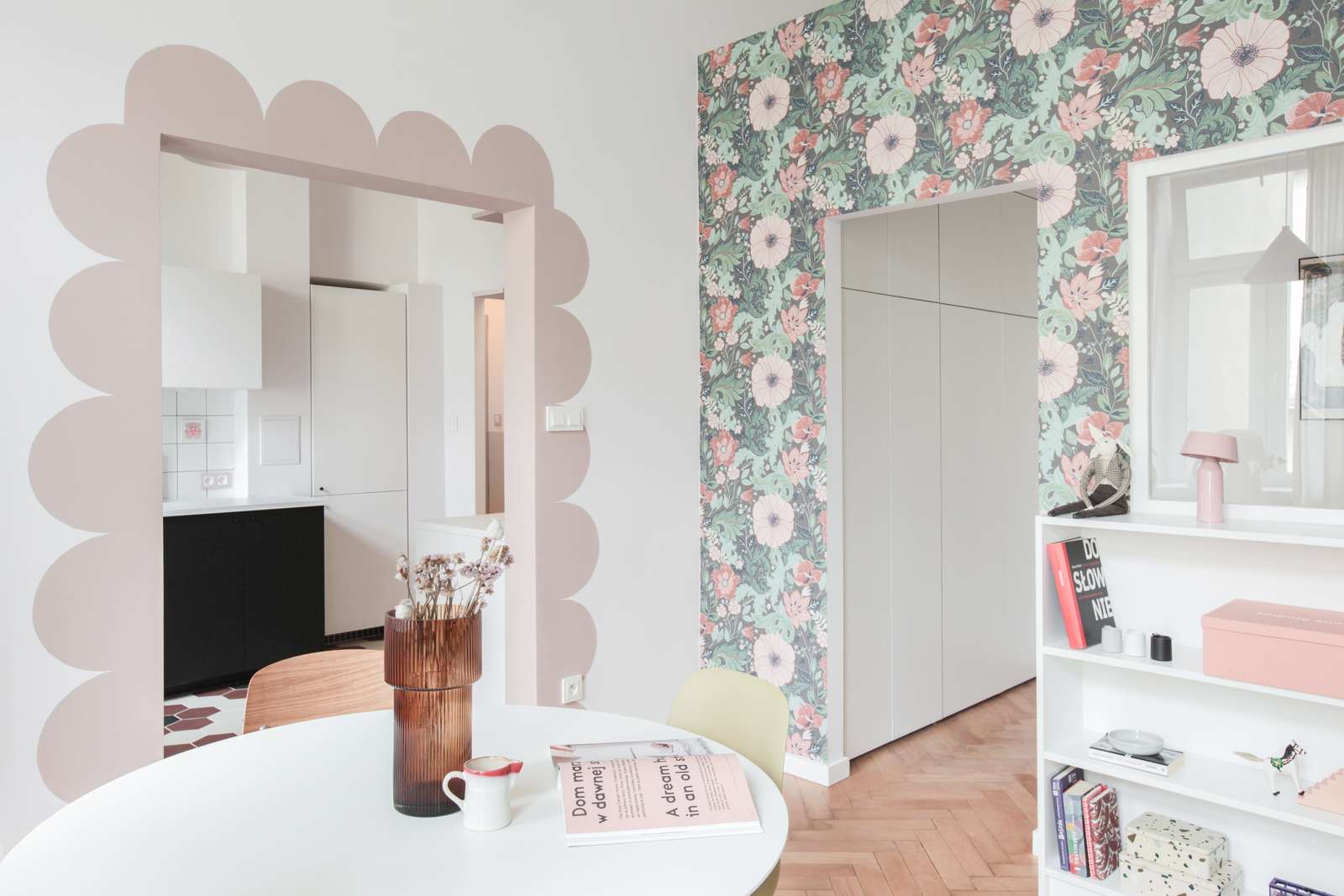 Bức tường chuyển tiếp giữa khu vực phòng bếp và phòng khách - phòng ăn được trang trí như những cánh hoa màu hồng phấn điệu đà trên tường. Một chiếc bàn ăn hình tròn được bố trí để phân vùng giữa phòng bếp và phòng khách.
