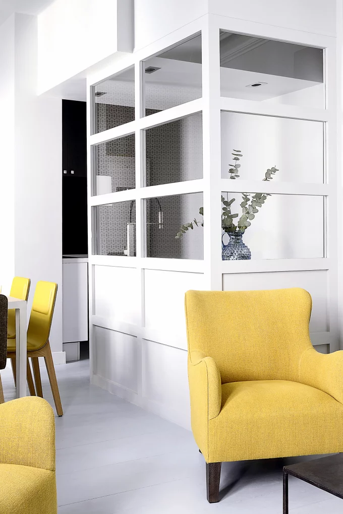 Phòng bếp siêu nhỏ chọn kiểu 'đóng khung' với một phần cửa kính bao quanh góc tạo nên 'ốc đảo' riêng tư. Phòng bếp tối giản, đơn sắc trở thành phông nền hoàn hảo cho những chiếc ghế màu vàng ở phòng khách và phòng ăn thêm nổi bật.