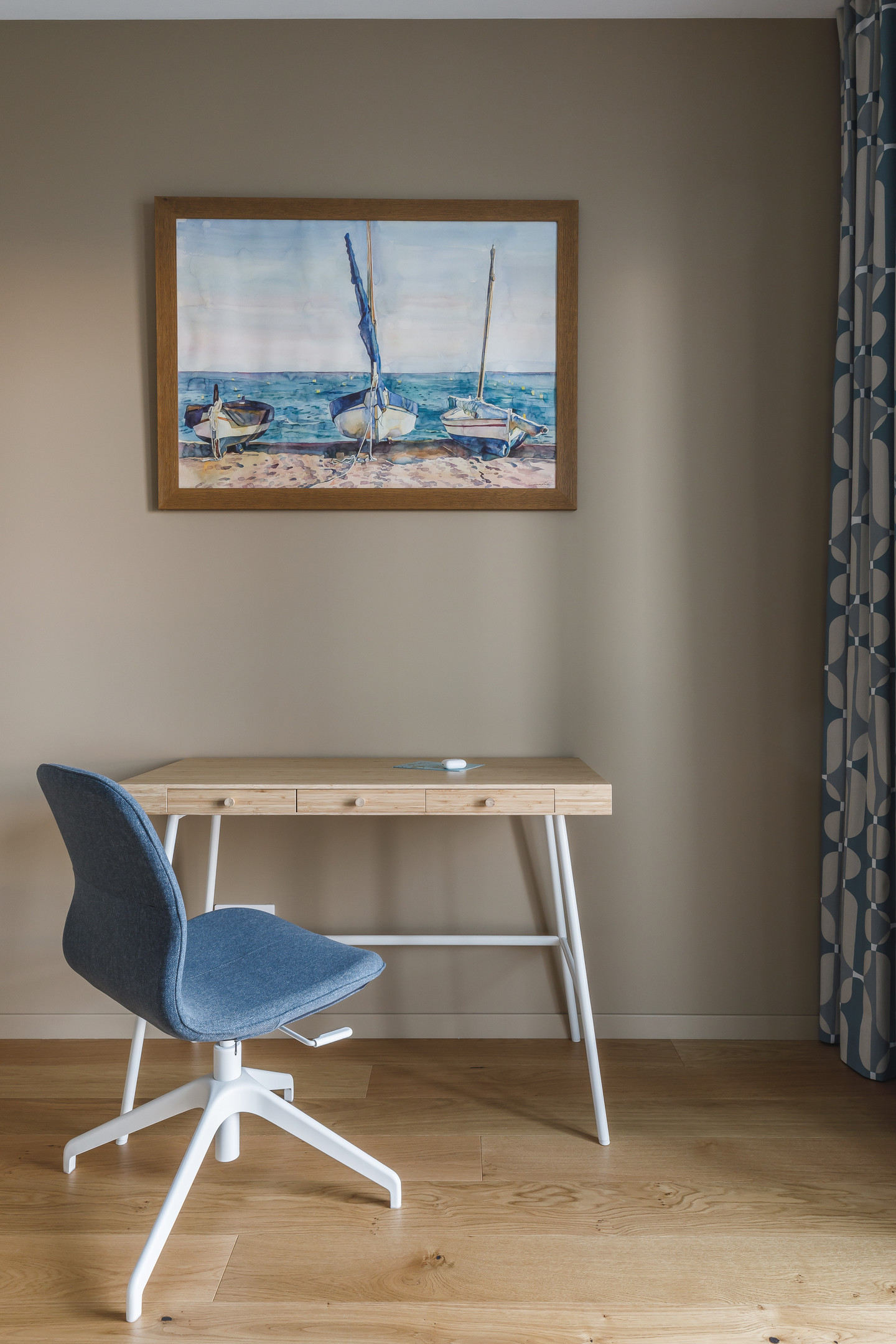 Đối diện giường ngủ là bộ bàn ghế tối giản với chiếc bàn gỗ và ghế ngồi màu xanh lam, trên tường là tranh treo vẽ cảnh biển sinh động.