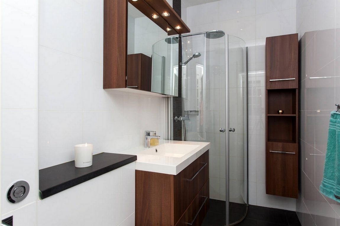 Phòng tắm sử dụng tone màu trắng sáng kết hợp nội thất gỗ tối màu để tạo sự ấm cúng nhất định. Buồng tắm đứng phân vùng với toilet bằng cửa kính trong suốt. Tủ lưu trữ gắn tường cửa gương soi '2 trong 1' tiện lợi.