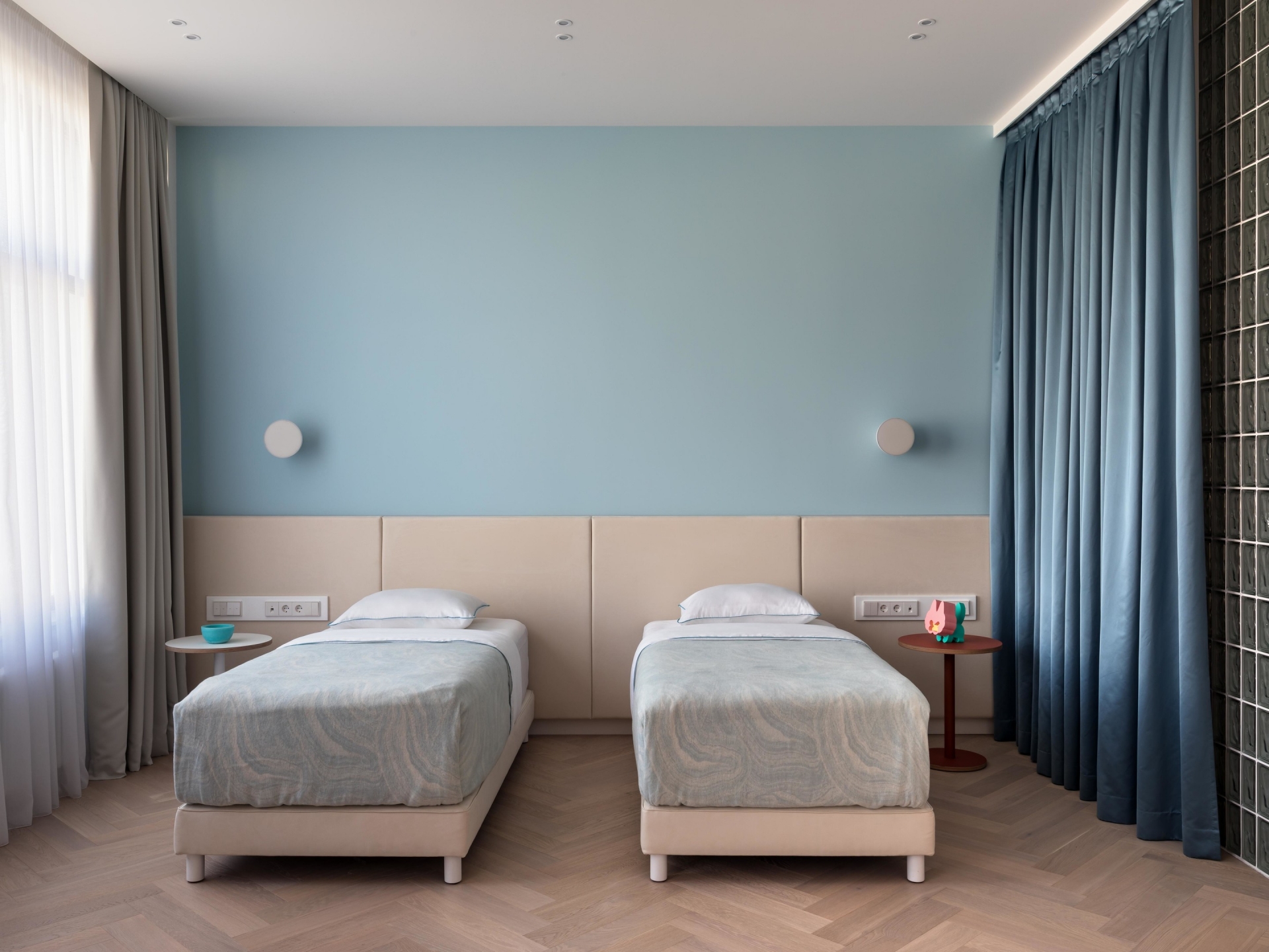 Phòng của hai bé trai đang tuổi tiểu học được thiết kế tối giản với 2 chiếc giường đơn bố trí song song, táp đầu giường và đèn gắn tường cũng nhỏ gọn. Tone màu chủ đạo là xanh lam nhẹ nhàng tươi mới.