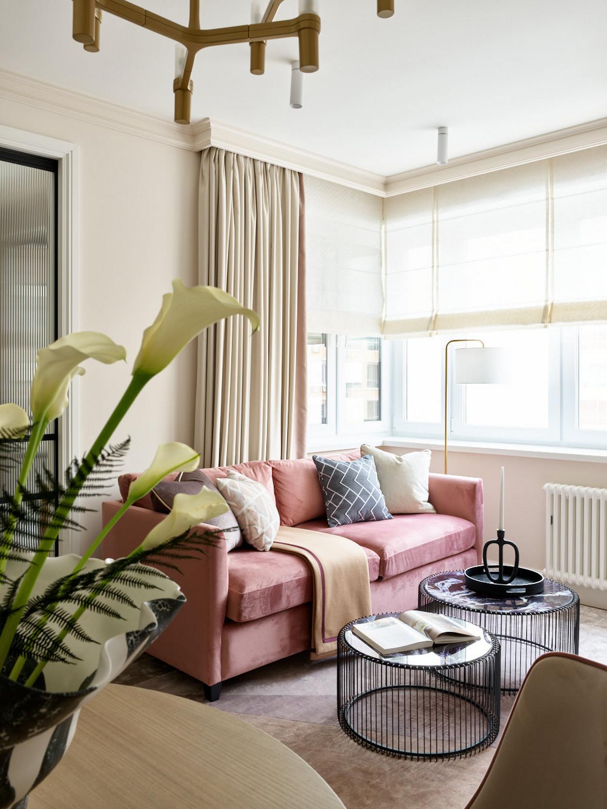 Phòng khách nhỏ xinh bên ô cửa đầy nắng ấm với ghế sofa màu hồng nổi bật giữa phông nền trung tính. Bộ đôi bàn nước thiết kế kiểu lồng chim, thêm chân nến trang trí cho vẻ đẹp cổ điển.