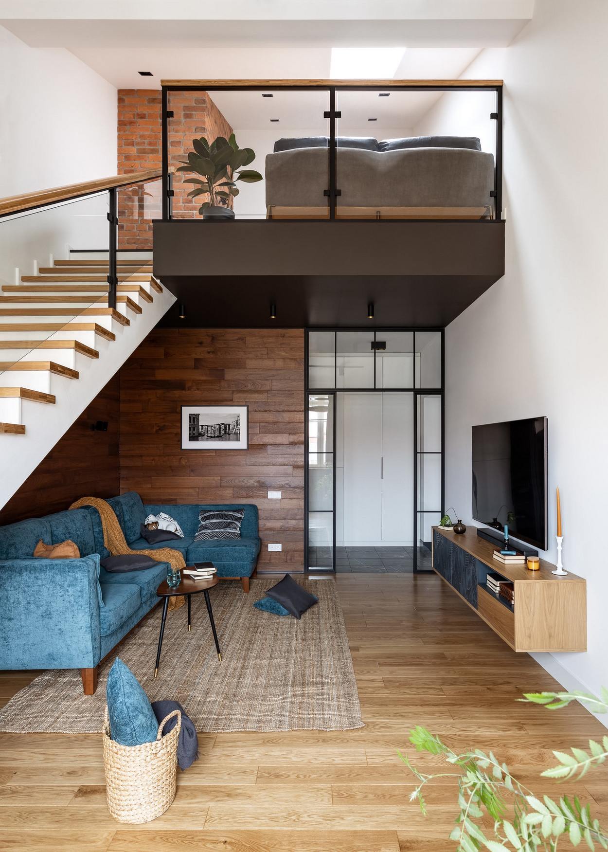 Phòng khách phân vùng với khu vực lối vào bằng cửa kính trượt khung màu đen thoáng sáng. Tường và sàn phòng khách sử dụng gỗ 2 tone màu đậm - nhạt để không gian sinh động hơn.