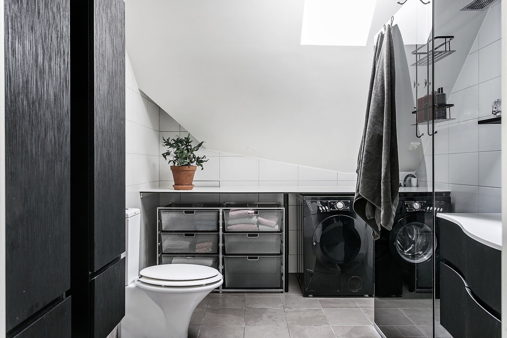 Khu vực nhà vệ sinh kết hợp phòng giặt ủi tiện lợi cho cả gia đình. Máy giặt và hệ tủ lưu trữ màu đen trở nên nổi bật giữa trần - tường trắng và sàn lát gạch xám.