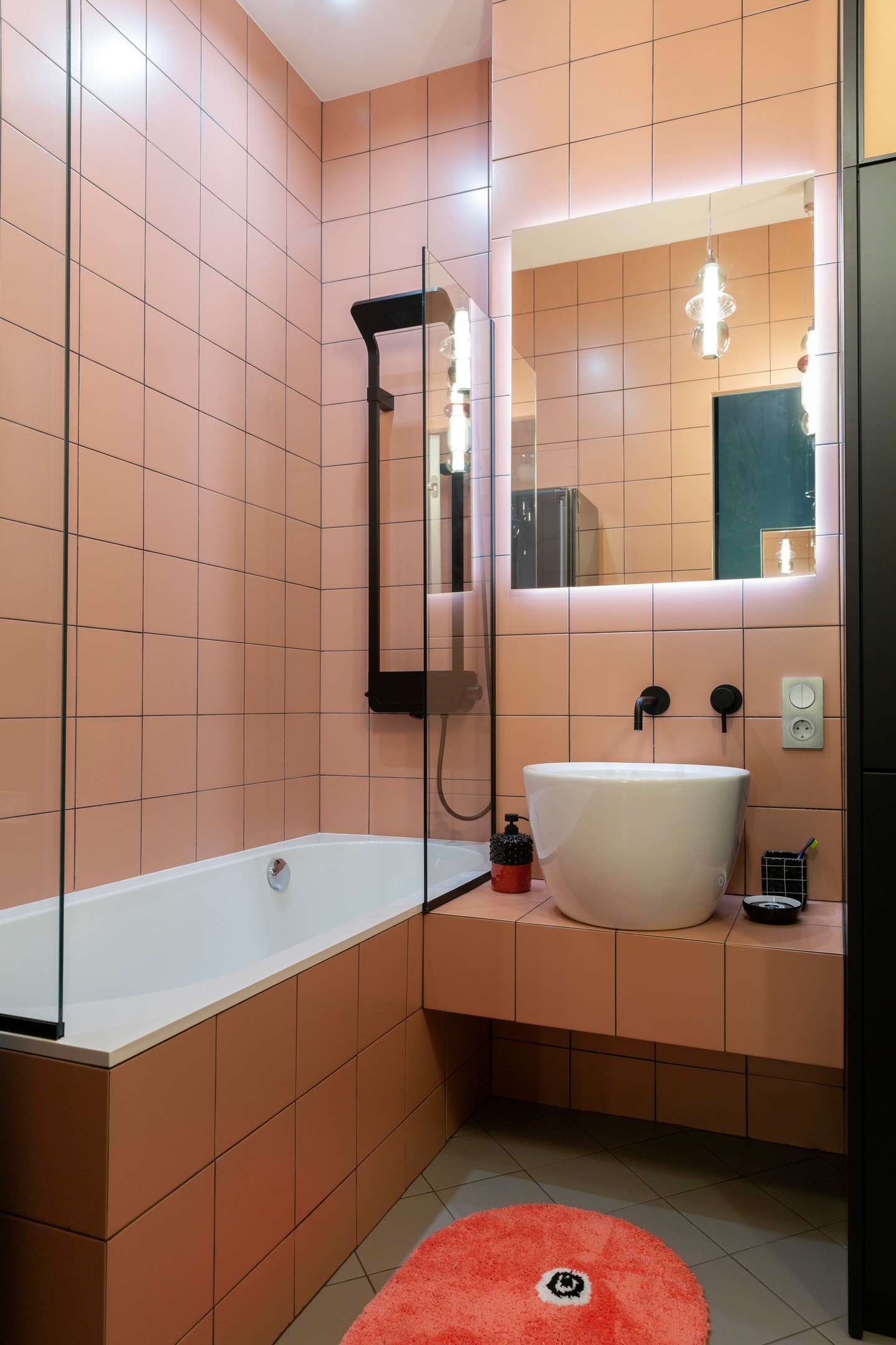 Khu vực bồn tắm nằm được phân vùng với nhà vệ sinh bằng cửa kính trong suốt, bên dưới sàn nhà bố trí một tấm thảm trải sàn hình oval màu cam đậm bắt mắt. Tấm gương tích hợp đèn Led trên tường bồn rửa cho căn phòng bừng sáng hơn!