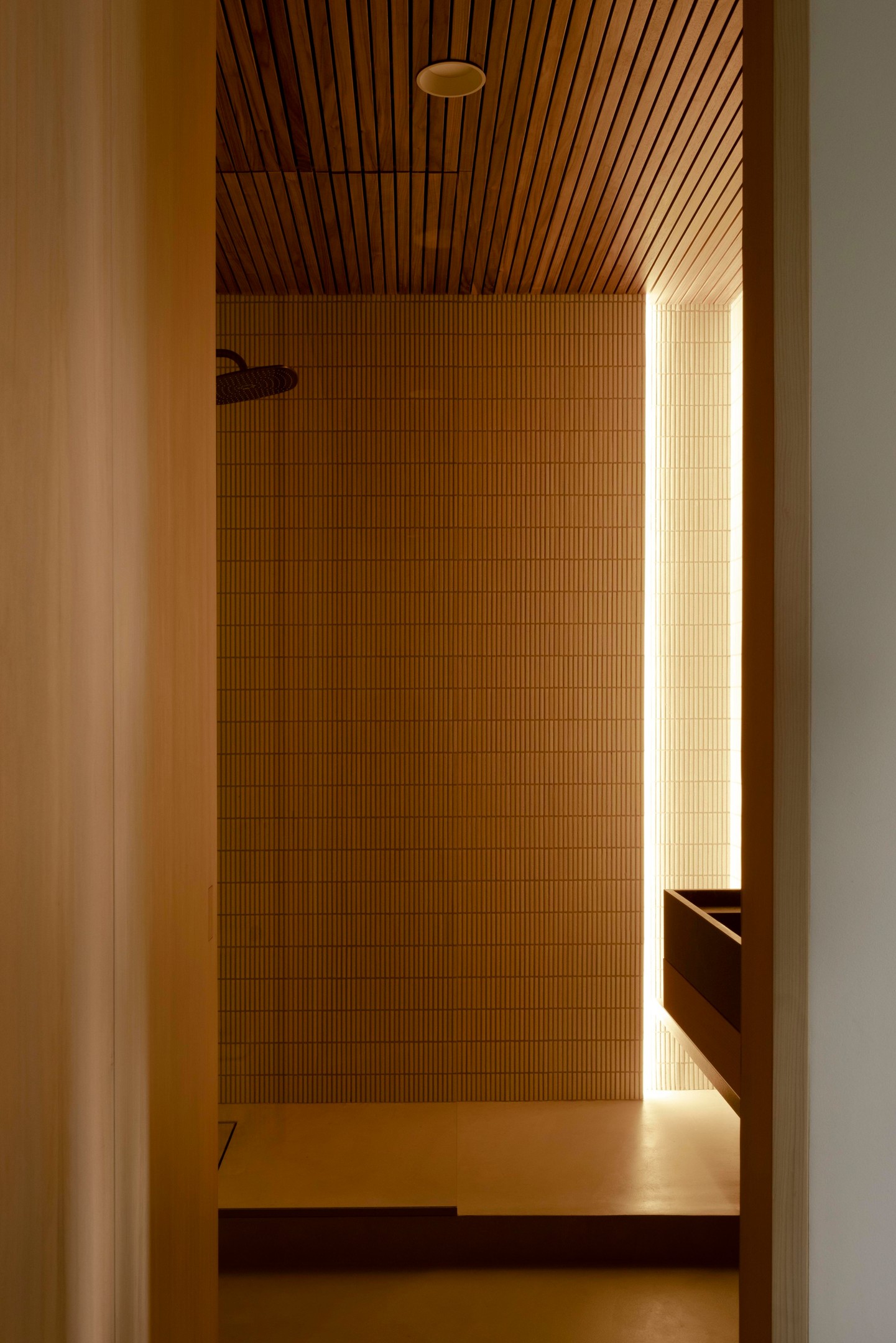 Bên trong, buồng tắm đứng vòi sen được phân vùng với nhà vệ sinh bằng vách ngăn kính trong suốt. Trần nhà ốp những thanh lam gỗ xuyên suốt từ hành lang cho cái nhìn ấm áp.