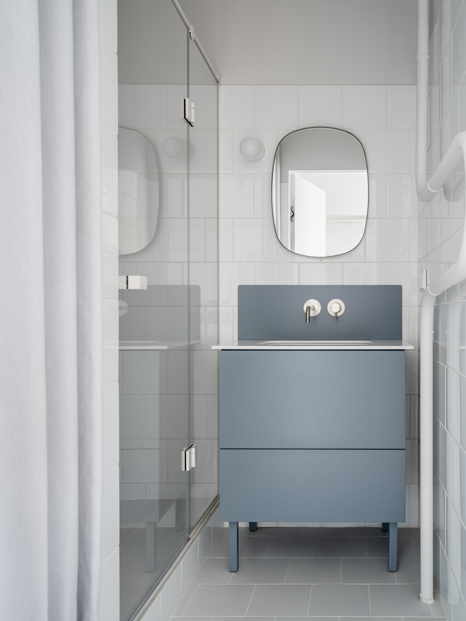 Phòng tắm nhẹ nhàng với chiếc tủ vanity màu xanh lam nhạt nổi bật trên nền tường ốp gạch trắng. Cửa kính trong suốt kết hợp rèm vải giúp phân vùng buồng tắm và nhà vệ sinh đẹp mắt.