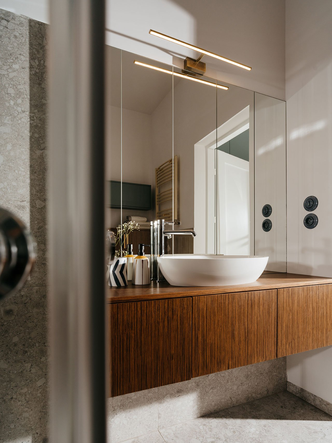 Phòng tắm sang chảnh với bức tường ốp gương cỡ lớn, phản chiếu không gian và ánh sáng tuyệt đẹp. Phía trên là chiếc đèn mạ vàng đồng cho ánh sáng ấm áp, kết hợp tủ vanity gỗ tone trầm.