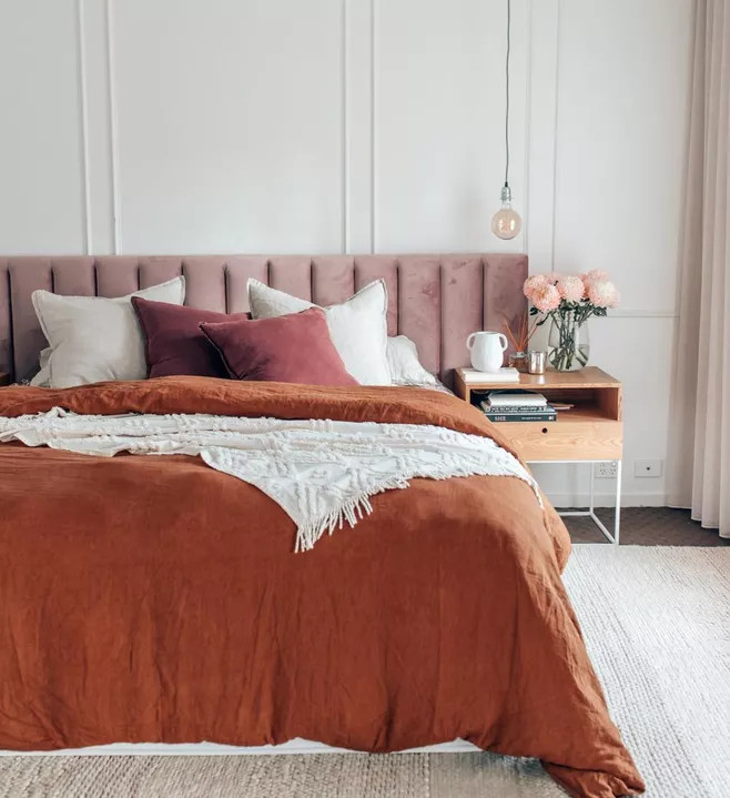 Phòng ngủ tinh tế với các gam màu hồng tím của vải nhung bọc đầu giường cùng bộ chăn ga gối màu cam cháy ấm áp, thêm vào lọ hoa với sắc hồng san hô là một điểm nhấn tuyệt vời.