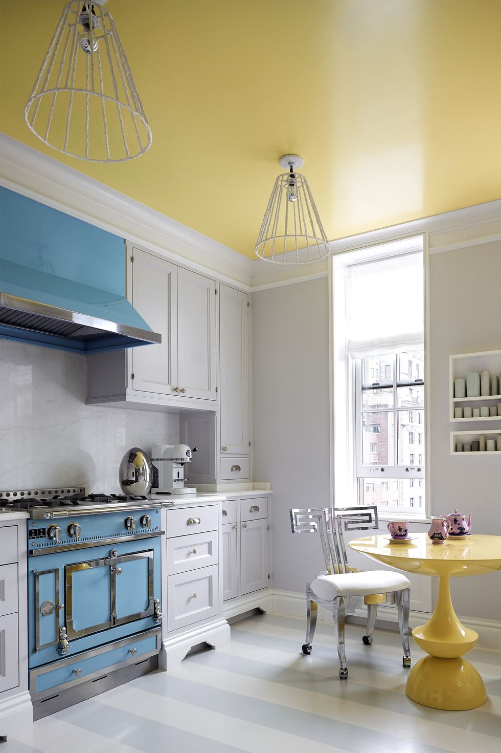 Không gian thiết kế theo phong cách vintage với tủ bếp, bàn ghế, đèn trần kiểu cổ điển. Sự tương phản giữa sắc vàng tươi của trần nhà, bàn ăn và tủ bếp xanh lam ở trung tâm, bao phủ bởi sắc trắng càng thêm phần nổi bật.