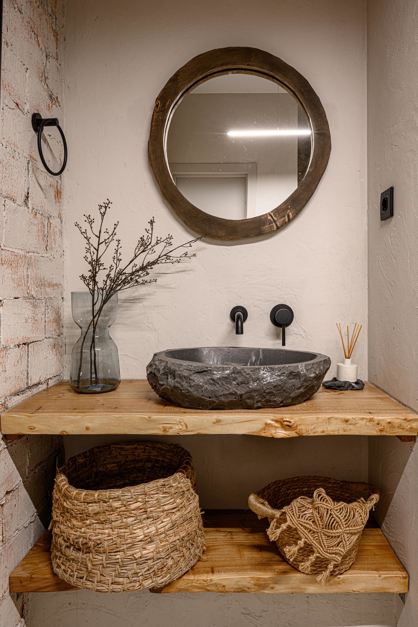 Một góc bồn rửa trong phòng tắm với những chiếc kệ được làm bằng gỗ tự nhiên, một phần tường ốp gạch thô, đặc biệt là chiếc bồn rửa bằng đá và những chiếc giỏ đan thô sơ mà quyến rũ.
